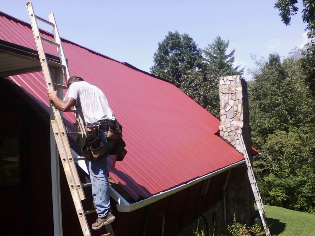 Roofer on a ladder beside a red metal roof and stone chimney — Soffit & Fascia Repair by Roofing Brothers in Nordman